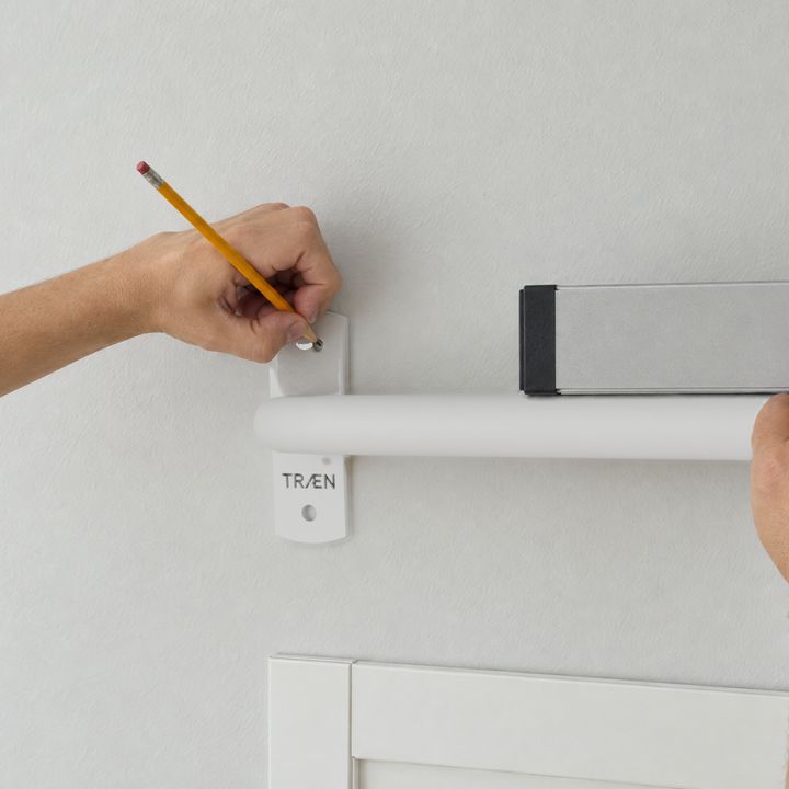 Zwei Hände halten die Stange über dem Türrahmen an die Wand; ein Stift zeichnet die Löcher an.